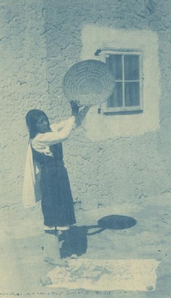 Pueblo Girl Winnowing Beans, girl with a Pima Indian basket from Arizona winnowing beans in front of an adobe structure by 
																			Charles F Lummis
